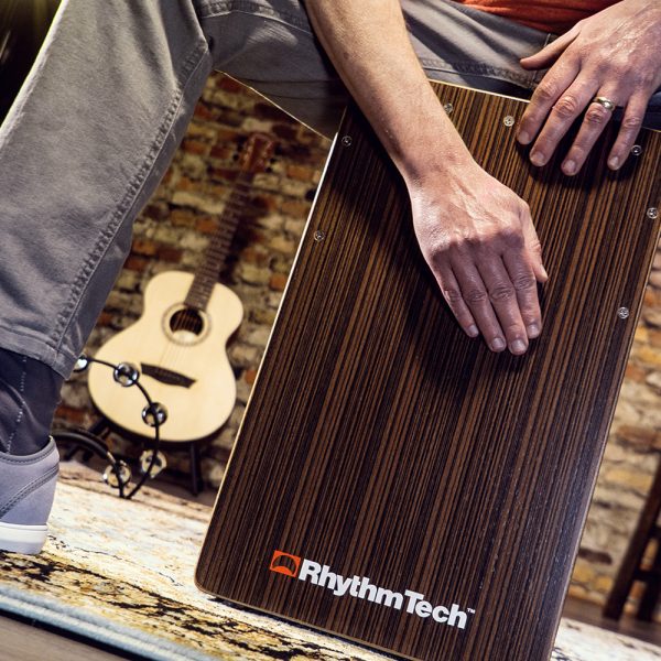 partial view of man playing Rhythm Tech cajon in front of acoustic guitar and brick wall