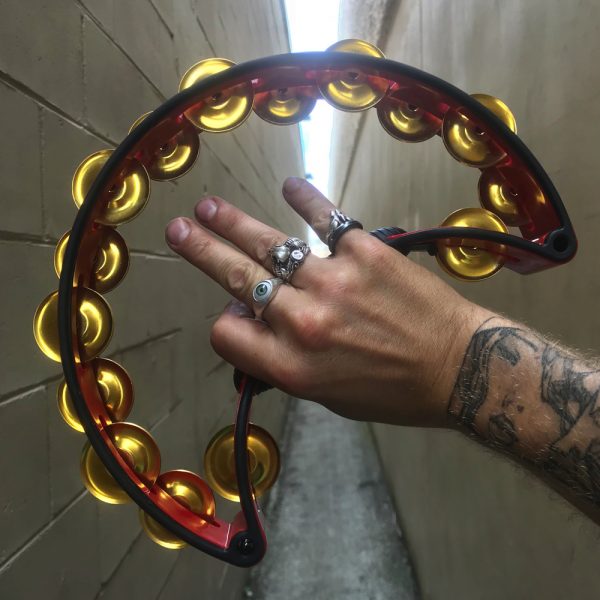 man's hand with tattoos and rings holding black Rhythm Tech tambourine in alleyway